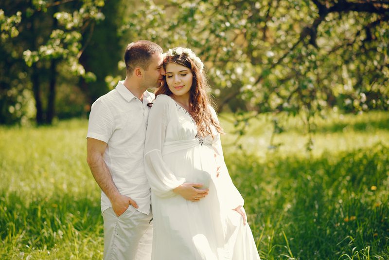 beautiful young pregnant girl in a long white dress and wreath on her head standing in a sunny summer park between the trees with her husband