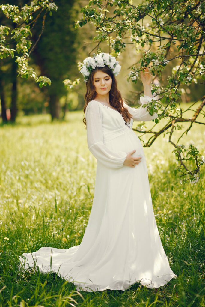 beautiful young pregnant girl in a long white dress and wreath on her head walking in a sunny summer park between the trees