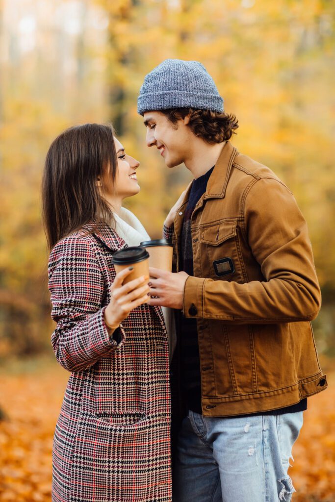 Couple in love with scarves sits in park holding cups of tea or coffee. Girl and bearded guy on a date hugging.