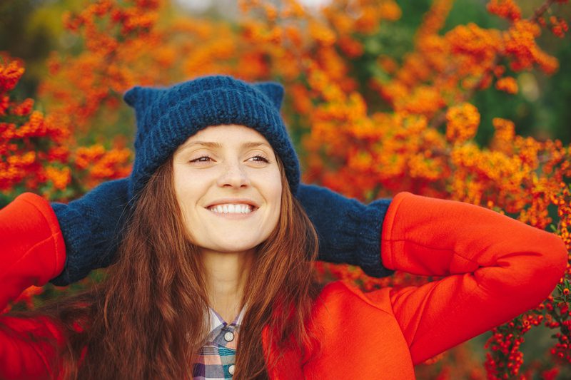 Fall senior photo ideas - Outdoor close up portrait of young beautiful happy smiling hipster girl posing on street, looking at camera. Model wearing stylish winter beanie hat, red coat, gloves. City lifestyle   