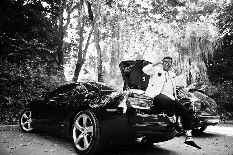 Stylish hipster man guy in sunglasses posed outdoor in street and sitting on the trunk his black muscle car. Rich black man.