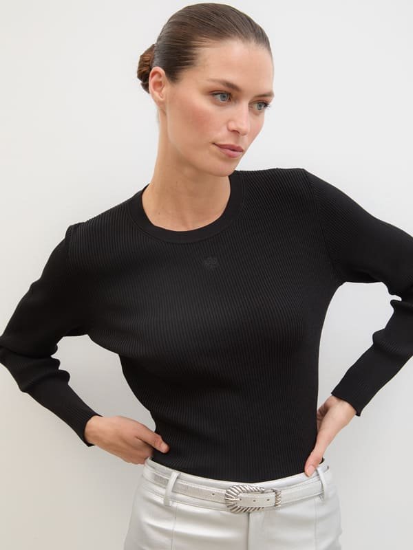Woman in black ribbed top with light pants and textured belt standing against plain background