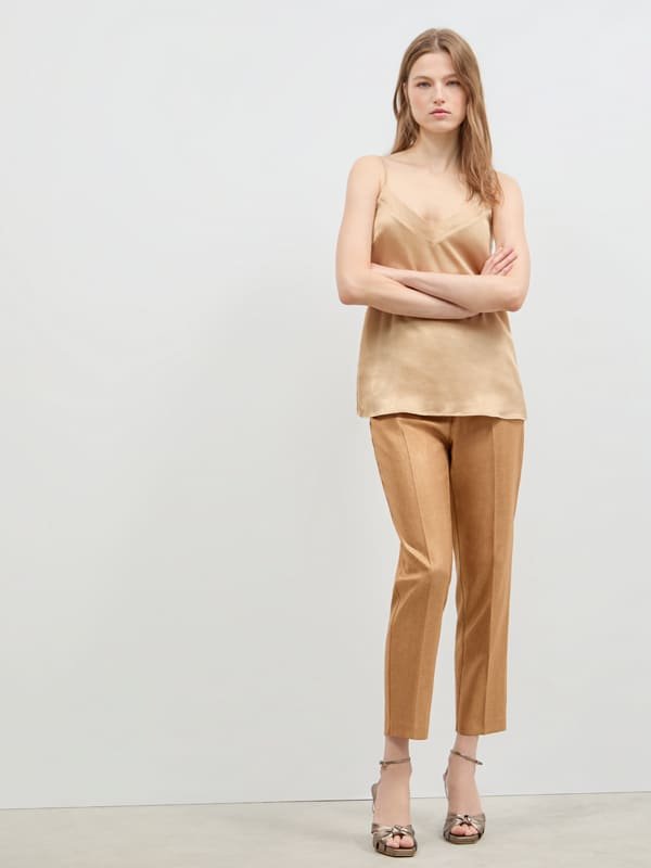 Woman wearing beige satin camisole, light brown tailored pants, and open-toed heels standing with arms crossed.