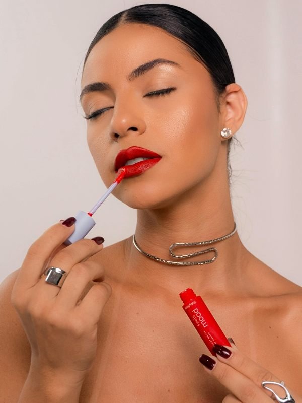 Woman with dark hair applying bright red lip gloss, wearing silver jewelry and dark nail polish.