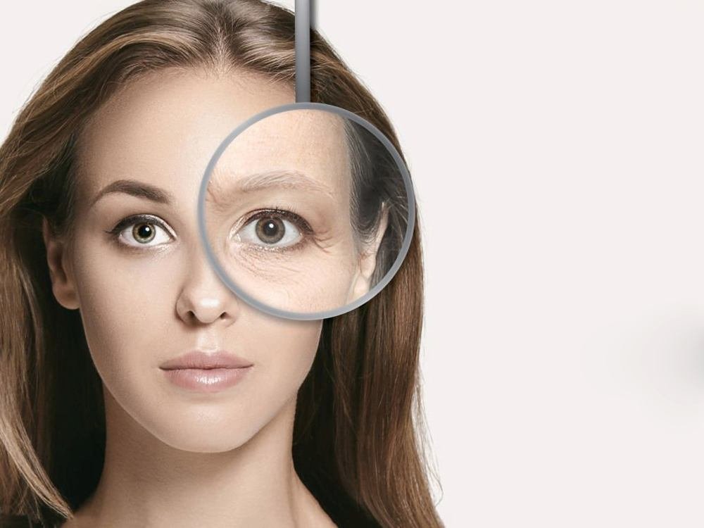Woman's face with magnifying glass showing comparison of youthful and aged skin around one eye