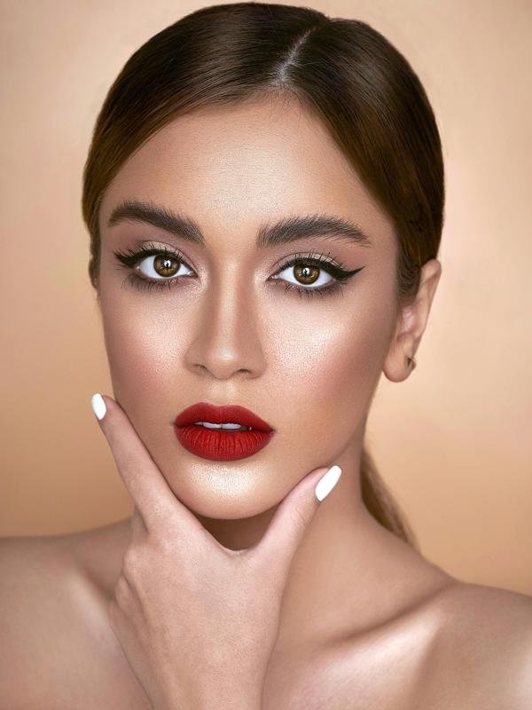 Close-up of a woman with red lipstick, winged eyeliner, white nail polish, and hand under chin.