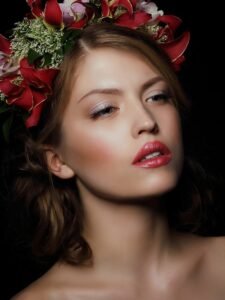 Close-up of a woman with wavy brown hair, floral headpiece, light makeup, and pinkish-red lipstick.