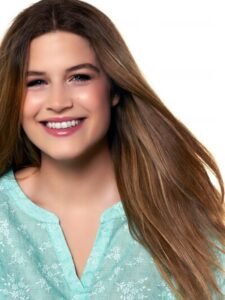 Young woman with long brown hair wearing a light blue floral top smiling against white background