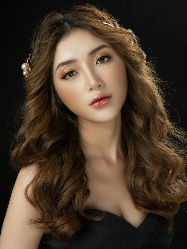 Young woman with wavy brown hair, decorative hairpins, warm makeup, and black strapless top on dark background.