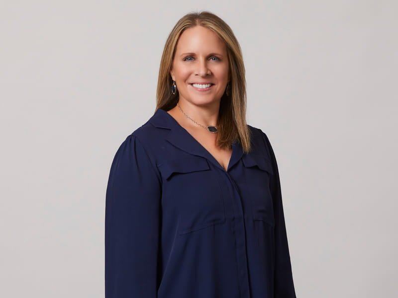 Smiling woman with shoulder-length light brown hair in a navy blouse against a light gray background.