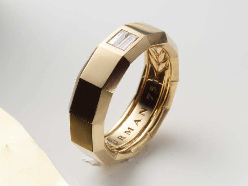 Gold-tone ring with geometric faceted band, baguette-cut diamond on outer top edge, inner engraving.