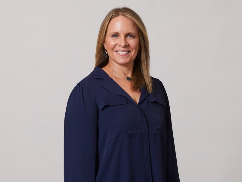 Smiling woman with shoulder-length light brown hair in a navy blouse against a light gray background.